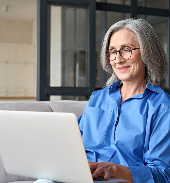 Older smiling 60s woman sitting at home on sofa, holding laptop.