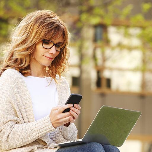 Portrait of busy sales woman sitting at business park and using her laptop while writing text on mobile.