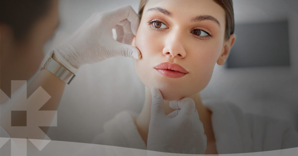 A woman undergoing an aesthetic or dermatologic consultation looks slightly away from the camera as a medical professional examines her face.