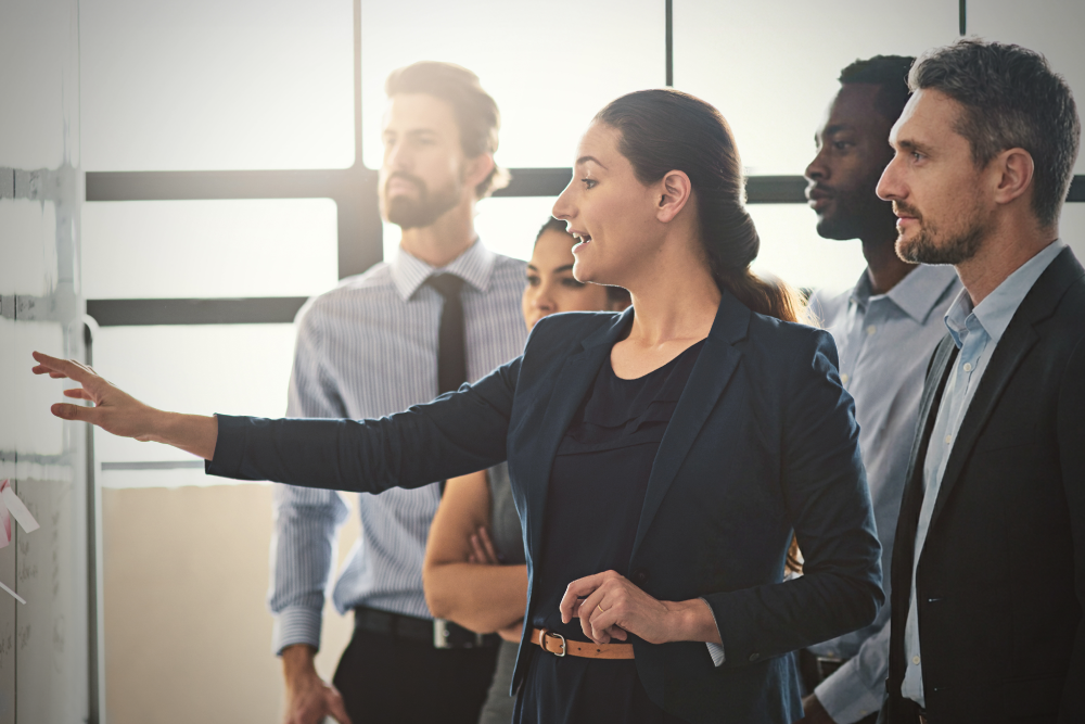 A business professional presents information on a whiteboard during a team meeting while several colleagues stand nearby and listen in a bright office setting.
