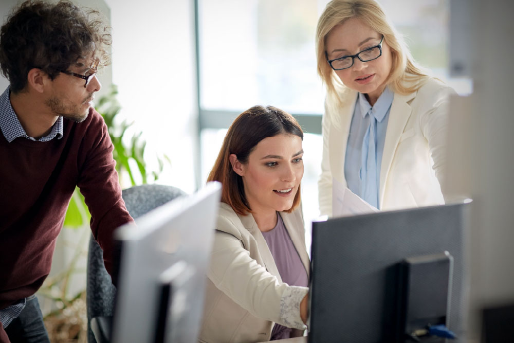 Three coworkers gathered around a computer monitor in an office, with a woman in a light blazer pointing at the screen while a man and another woman look on attentively.
