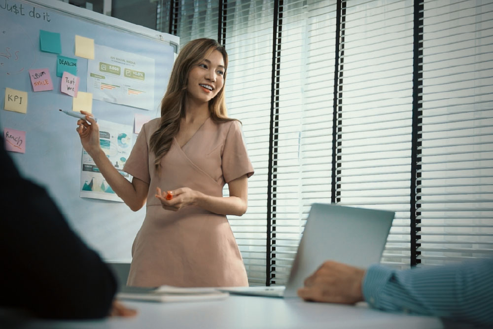 Woman giving a presentation in a modern office, standing beside a whiteboard with charts and notes.