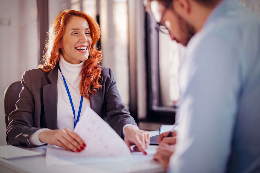A smiling woman with red hair and a lanyard sitting at a desk, holding a document and reviewing it with a colleague who is writing in the foreground.